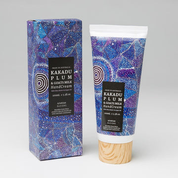 Kakadu Plum hand cream 100ml, white plastic tube has wooden lid; giftbox. Artwork by Warlukurlangu artist Alma Granites. Background vibrant blue and purple; black circles; white dot art. Australian made.