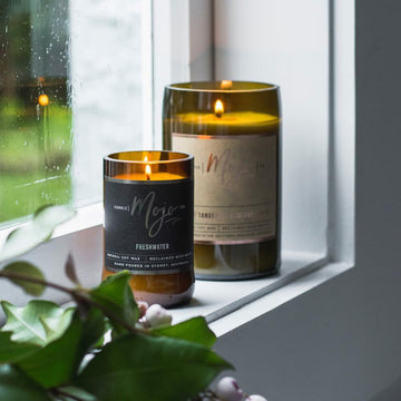 A candle made from a wine bottle with a cork lid is lying down surrounded by flowers and spices including a rose and lavender.