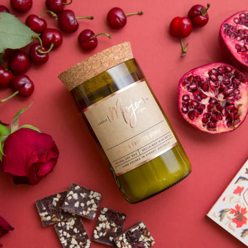 A candle made from a wine bottle with a cork lid is laying down surrouned by cherries, chocolate and pomegrantes.
