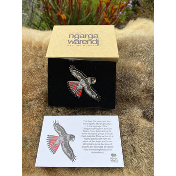 Enamel on metal brooch of a black and white patterned red tailed black cockatoo. The black cockatoo has black featured on the head and wings and is in flight position. 