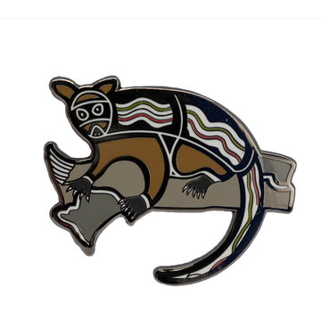 Enamel on metal brooch of a black, white and brown patterned possum. The possum has black featured around the head and body and is in a sitting position on a grey coloured tree branch. 