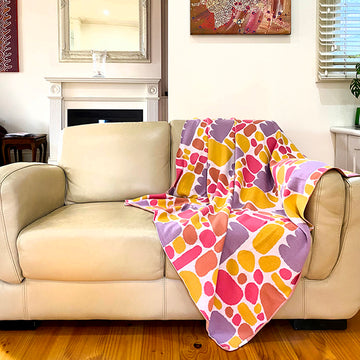 A throw is draped over a couch. The throw features artwork by Keturah Nangala Zimran in yellow and purple.