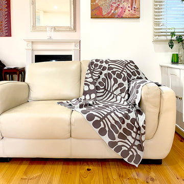 A throw is shown draped over a couch. The throw features artwork by Mitjili Napurrula in brown and white.