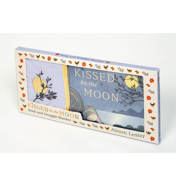 A box with a large clear window on the front showing the 'Kissed by the Moon' book inside and a folded up blanket. Both items feature the moon shining brightly behind a tree branch.