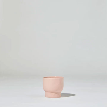 A small pink ceramic cup with a tapered base is sitting against a grey background.