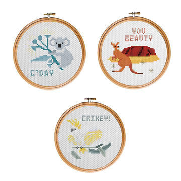 Three cross stitch designs inside wooden round hoops. The left design is a koala in a tree with the text 'G'Day'. The right is a kangaroo standing in front of Uluru with the text 'You Beauty'. The bottom design is a cockatoo on a branch with the text 'Crikey!'.