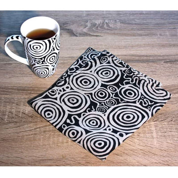 A linen tea towel printed with a black and white circle design by Nelly Patterson is laying folded up on a wooden table next to a mug with the same pattern.
