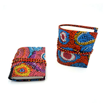 Two small notebooks pictured with red rope around them and printed with artwork by Andrea Mimpitja Adamson on the covers. One notebook is lying flat and the other is standing up.