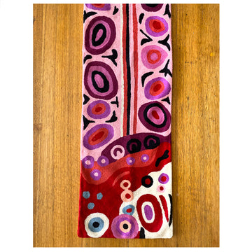 A table runner made from wool with a chain stitch design of Andrea Mimpitja Adamson's artwork in purple and red. The table runner is laying on a table and the photo is a birds eye view.