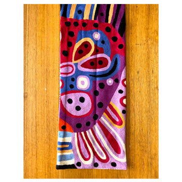 A table runner made from wool with a chain stitch design of Andrea Mimpitka Adamson's artwork in purple, blue and red. The table runner is on a wooden table and the photo is a birds eye view.