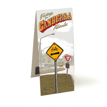 A magnetic bookmark standing open and printed with artwork by Trevor Dickinson of a street sign with a bike and arrow on it. The heading 'Greetings from Canberra Australia' is at the top.