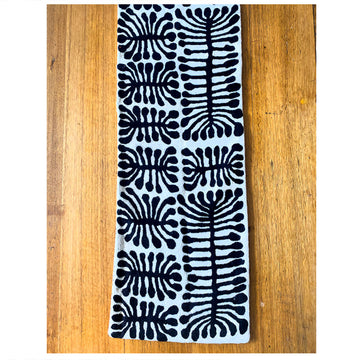 A birds eye view of a wool chain stitched table runner on a wooden table. The table runner features artwork by Mitjili Napurrula in black and white.