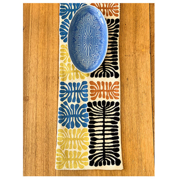 A birds eye view of a wool chain stitched table runner on a wooden table with a oval shaped blue bowl on top. The table runner features artwork by Mitjili Napurrula in blue, black, orange and yellow.