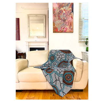 A cotton throw is on a cream coloured couch inside a house with artwork on the wall behind. The throw is printed with artwork by Bianca Gardiner-Dodd in blue and orange dots.
