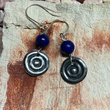A pair of drop earrings with a blue gemstone and sterling silver circle with circular indents against a sandstone rock.
