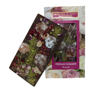 A block of chocolate is leaning against its packaging box. The chocolate has turkish delight, pistachios and rose petals attached to it and the box is printed with flowers with a window in the middle showing the chocolate inside.