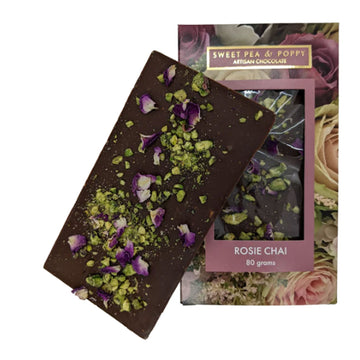A block of chocolate is leaning against its packaging box. The chocolate has pistachios and rose petals attached to it and the box is printed with flowers with a window in the middle showing the chocolate inside.