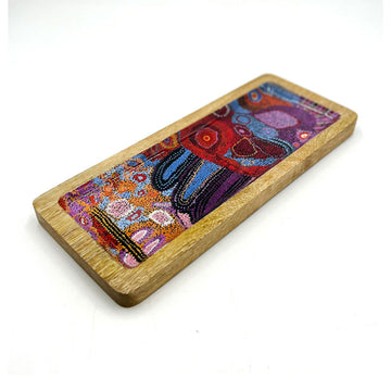 A wooden rectangle tray on an angle printed with artwork by Andrea Mimpitja Adamson inside.
