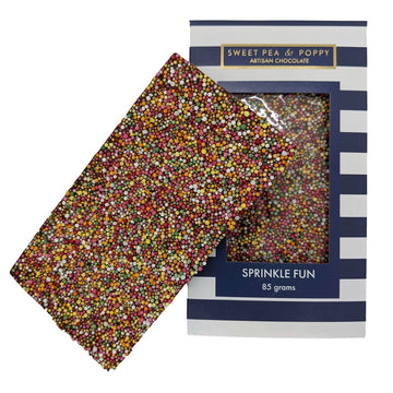 A block of chocolate is leaning against its packaging box. The chocolate has coloured sprinkles attached to it and the box is printed with flowers with a window in the middle showing the chocolate inside.
