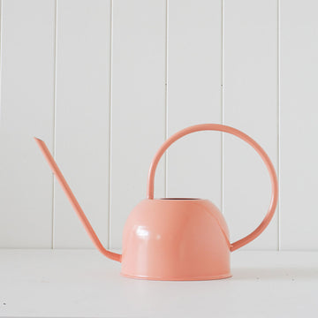 A watering can with a big round handle in peach against a white panelled wall.