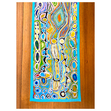 A top down view of a wooden table with a table runner laying flat on it. The runner features artwork by Judy Watson in blue, yellow and black.