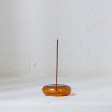 An amber coloured glass incense holder with a round shape and an incense stick vertical in the middle.