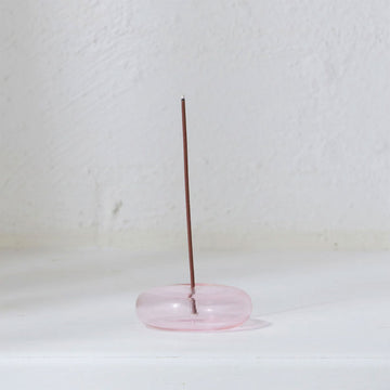 A pink glass incense holder in a round shape with a stick vertically inside.