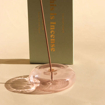 A pink glass incense holder in a round shape with a stick vertically inside.