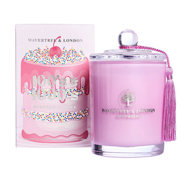 A glass candle filled with pink wax and a tassel on the lid. There is a box next to the candle illustrated with a tall pink cake decorated with 100s and 1000s.
