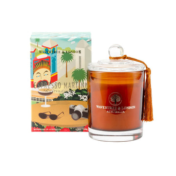 A glass container filled with golden brown candle wax and a tassel on the lid. The candle is next to a box with an illustration of a martini glass, sunglasses and camera on a table overlooking Luna Park.