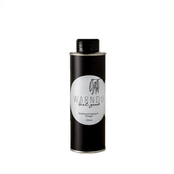 A black tall canister with a black lid and white round label on the front with the text 'Warndu Feel Good Wattleseed Balsamic Vinegar'.