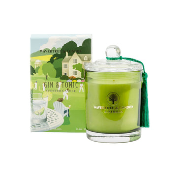 A clear glass container filled with green candle wax and a tassel hanging off the lid. The candle is next to a box illustrated with a glass of gin and tonic on a table overlooking people playing cricket.