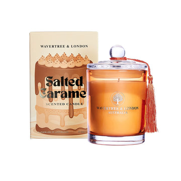 A glass container filled with caramel coloured candle wax and with a tassel on the lid. There is a box next to the candle illustrated with a caramel coloured cake.