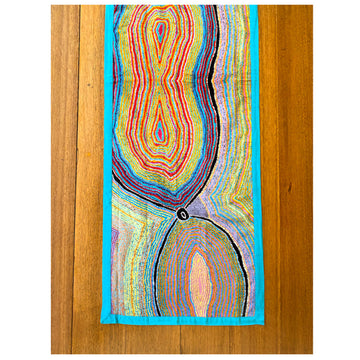 A top down view of a wooden table with a table runner on it. The runner features artwork by Liddy Napanangka Walker in blue, red and yellow.