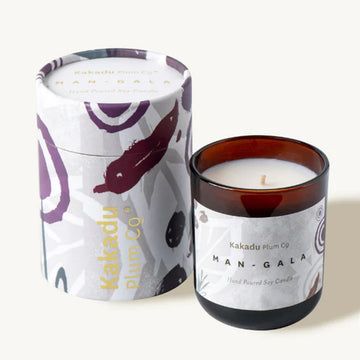 A glass candle next to its cylinder container. The container and candle are both printed with a different design and have the text 'Kakadu Plum Co Mangala' written on them.