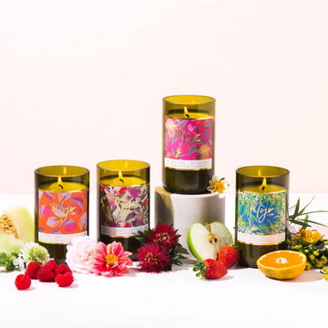 A candle made from a recycled wine bottle with a cork lid and a floral printed product label. There are fruits and flowers surrounding the candle.