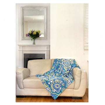 Indigenous art cotton throw, featuring artwork by Cedric Varcoe, flung over a white leather sofa. White painted walls, fireplace, vase of yellow flowers on the mantelpiece.