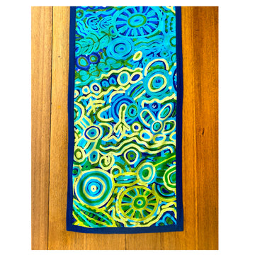 Top down view of a wooden table with a table runner on top. The runner features artwork by Cedric Varcoe in blues and yellows.