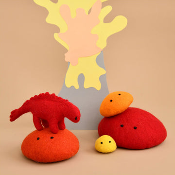 A red felt dinosaur toy is standing on top of an orange felt rock. There are three different sized felt rocks to the right and a volcano made from paper at the back.