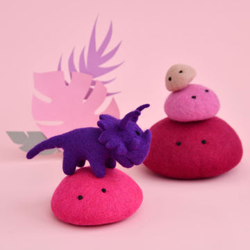 A purple triceratops dinosaur toy made from felt is standing on a pink felt rock. There are three stacked felt rocks in the background as well as paper leaves.