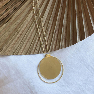 A round gold necklace with a hollow circle outline hanging off a gold chain over a palm leaf.