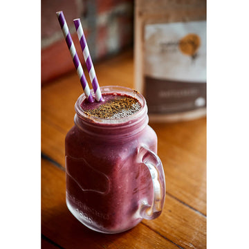 Amber coloured glass jar, black plastic lid, label with indigenous motif. Kakadu Plum Co, Australian Sustainable, wattleseed ground, type printed on label. Logo printed on tamper proof tab.Thick red smoothy served in in a chilled, handled jar, glass. Two purple and white cardboard straws, garnish on top. Smoothy is on a wooden bench, wattleseed packet at back.
