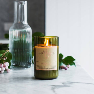A candle made from a wine bottle with a cork lid is lying down surrounded by fruits including watermelon, berries and lemons.