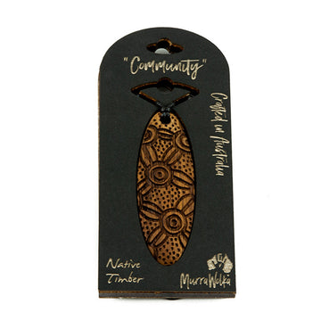 A wooden necklace with carvings is pictured inside it's cardboard packaging. The box features the words 'Community', 'Crafted in Australia', 'Native Timber' and 'MurraWolka'