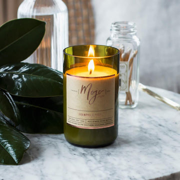 A candle made from a wine bottle is lying down on a white bench. Surrounding the cadle is a pen and notebook, books, flowers and nuts.