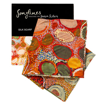 Silk scarf printed with the design 'Minyipuru at Pangkai' featuring green ovals with red outlines and orange and purple dots. The packaging slip is behind the scarf.