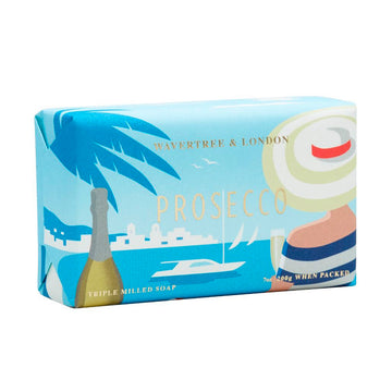Packaging of a bar of soap featuring an illustration of a person's back wearing a striped hat and striped shirt holding a glass of champagne. There is a champagne bottle on the left and a view of a boat in the water in the background. The word 'Prosecco' is written in gold across the middle.