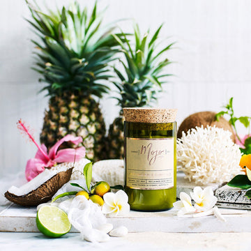 A candle made from a wine bottle with a cork lide is lying amount fruits including a mango and watermelon.