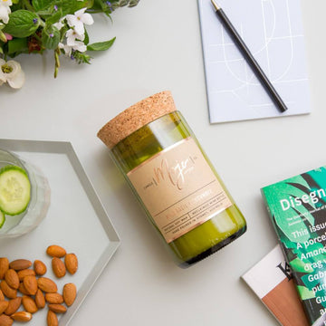 A candle made from a wine bottle is lying down on a white bench. Surrounding the cadle is a pen and notebook, books, flowers and nuts.